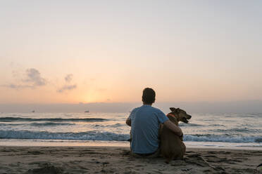Mann sitzt mit seinem Hund und genießt die Morgendämmerung am Strand - EGAF00585