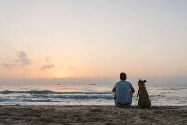 Mann mit seinem Hund am Strand sitzend in der Morgendämmerung - EGAF00586