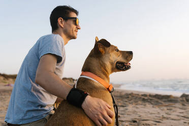 Mann und Hund schauen weg, während sie am Strand sitzen - EGAF00596