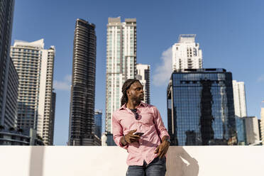 Nachdenklicher Afro-Mann mit Smartphone vor Wolkenkratzern in Miami, Florida, USA - MAUF03492