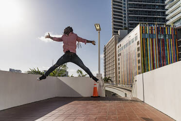 Fröhlicher Afro-Mann mit ausgestreckten Armen, der auf dem Fußweg gegen den Himmel springt, Miami, Florida, USA - MAUF03493