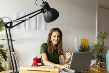 Lächelnde Geschäftsfrau mit Brille am Schreibtisch sitzend im Heimbüro - VABF03208
