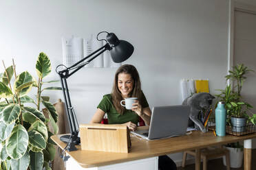 Lächelnde Unternehmerin, die eine Tasse hält und ein digitales Tablet auf dem Schreibtisch im Heimbüro benutzt - VABF03211
