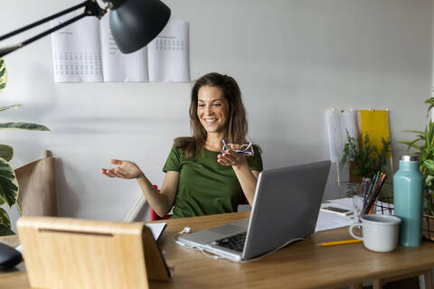 Smiling young woman gesturing while looking at digital tablet on desk in home office stock photo