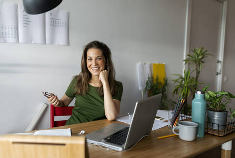 Smiling businesswoman sitting at desk against wall in home office stock photo