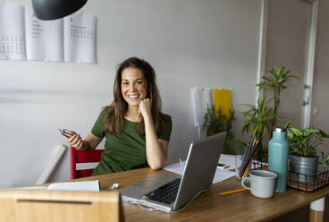 Smiling businesswoman sitting at desk against wall in home office - VABF03213
