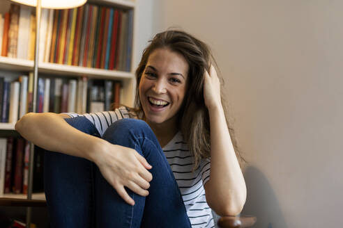 Fröhliche junge Frau mit Hand im Haar, die zu Hause an der Wand sitzt - VABF03226
