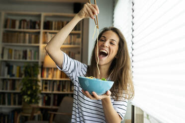 Junge Frau isst Spaghetti, während sie zu Hause am Fenster sitzt - VABF03231