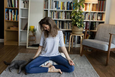 Happy young woman with cat sitting on rug against bookshelf at home - VABF03234