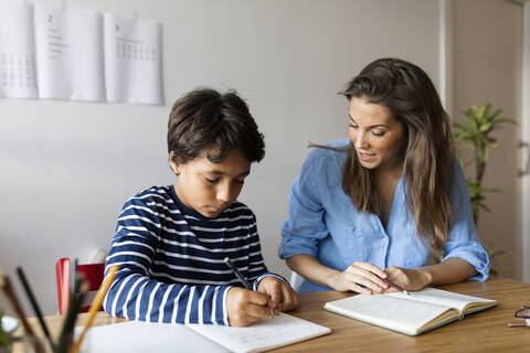 Female tutor assisting boy in writing homework on table at home stock photo