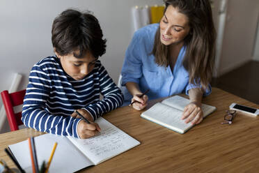 Young woman assisting boy in writing homework on table at home - VABF03251