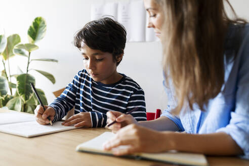 Nahaufnahme einer Nachhilfelehrerin, die einen Jungen beim Schreiben von Hausaufgaben auf dem Tisch beobachtet - VABF03253