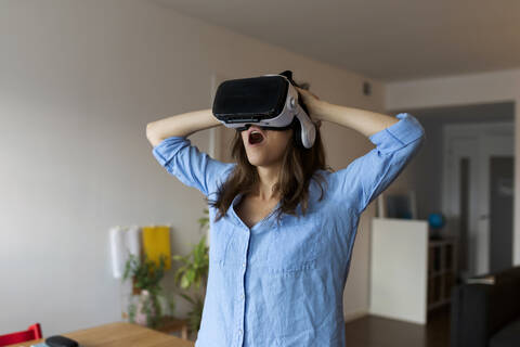 Young woman with mouth open looking through virtual reality simulator while standing in home office stock photo