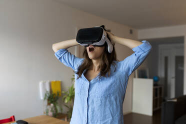 Young woman with mouth open looking through virtual reality simulator while standing in home office - VABF03257