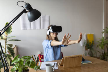 Young woman looking through virtual reality simulator gesturing while sitting in home office - VABF03259