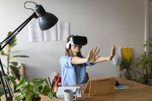 Junge Frau, die durch einen Virtual-Reality-Simulator schaut und gestikuliert, während sie im Büro zu Hause sitzt - VABF03259