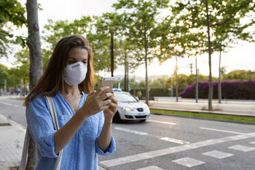 Young woman wearing mask using smart phone while standing on street in city - VABF03260