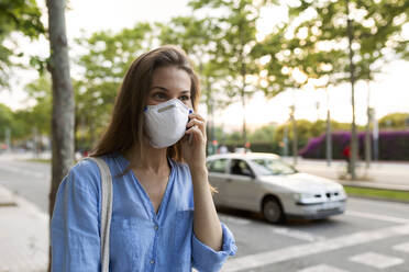 Young woman wearing mask talking over smart phone while standing on street in city - VABF03262