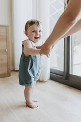 Baby girl holding mother's hands while learning to walk on floor at home - GMLF00357