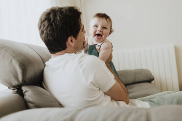 Father holding cheerful baby daughter while relaxing on sofa at home - GMLF00358