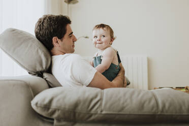 Father holding cute baby daughter while relaxing on sofa at home - GMLF00359