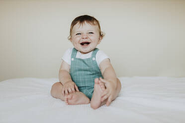 Cute baby girl laughing while sitting on bed against wall at home - GMLF00361