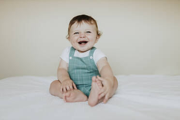 Cute Baby Mädchen lachen, während auf dem Bett gegen die Wand zu Hause sitzen - GMLF00361