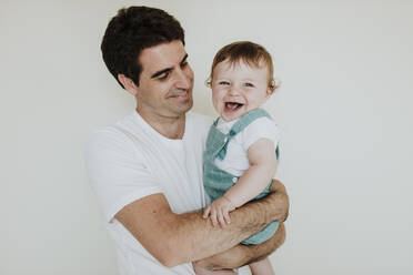 Father looking at cute cheerful daughter while carrying her against white wall - GMLF00367