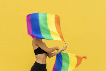 Woman waving rainbow flags while standing against yellow wall in city - VABF03270