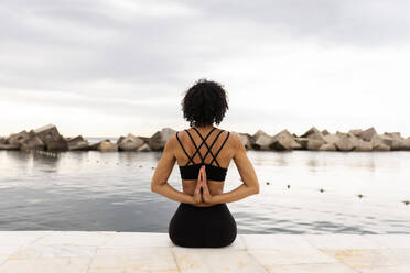 Mid adult woman with curly hair practicing yoga while sitting on promenade by sea - VABF03273