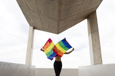 Mid adult woman waving rainbow flags while standing on built structure against sky - VABF03286