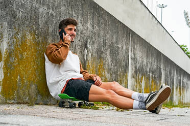 Junger lässiger Mann sitzt im Freien auf einem Skateboard und benutzt ein Telefon - CAVF87483