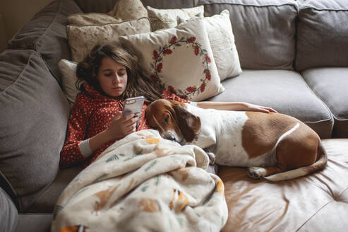 Tween-Mädchen schaut auf ihr Handy, während sie mit einem Hund auf der Couch kuschelt - CAVF87565