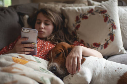 Tween-Mädchen schaut auf ihr Handy, während sie mit einem Hund auf der Couch kuschelt - CAVF87566