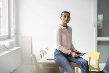 Portrait of businesswoman sitting on desk in loft office holding tablet - KNSF08226