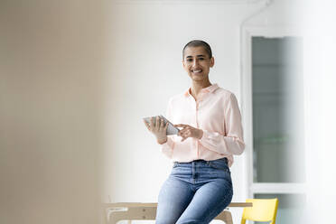 Portrait of smiling businesswoman sitting on desk in loft office holding tablet - KNSF08231