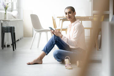 Portrait of woman sitting on the floor in a loft holding smartphone - KNSF08249