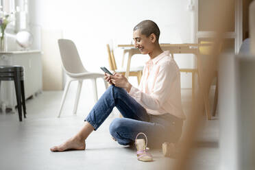 Happy woman sitting on the floor in a loft using smartphone - KNSF08250