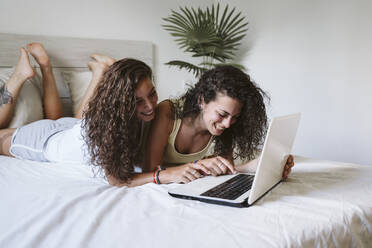 Glückliches lesbisches junges Paar, das einen Laptop benutzt, während es zu Hause auf dem Bett liegt - EBBF00459