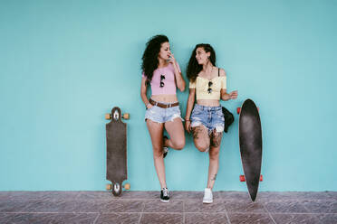 Lesbisches Paar, das raucht, während es mit Skateboards an einer blauen Wand in der Stadt steht - EBBF00474