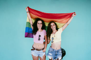 Cheerful lesbian couple holding rainbow flag while standing against wall - EBBF00484