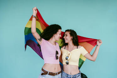 Happy lesbian couple holding rainbow flag looking at each other while standing against wall - EBBF00487