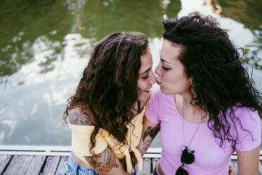 Nahaufnahme einer jungen Frau, die auf die Nase ihrer Freundin küsst, während sie am See sitzt - EBBF00498