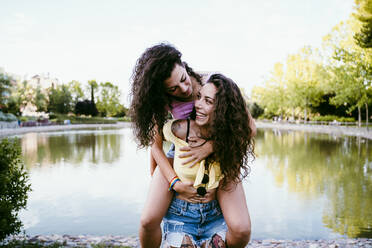 Cheerful young woman piggybacking girlfriend while standing against lake - EBBF00500