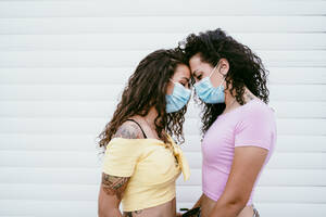 Lesbian couple wearing masks with face to face standing against wall in city - EBBF00506