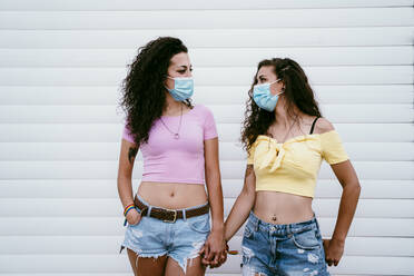Lesbian couple wearing masks holding hands while standing against wall in city - EBBF00508
