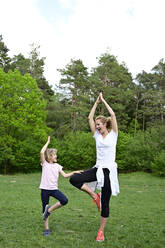 Happy mother and daughter practicing tree pose on grassy land in forest - ECPF00980