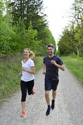 Smiling couple running on dirt road against trees and plants in forest - ECPF00998