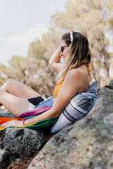 Junge Frau hört Musik über Kopfhörer, während sie mit einer Regenbogenflagge auf einem Felsen sitzt - MRRF00161