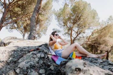 Junge Frau mit Sonnenbrille und Regenbogenflagge entspannt sich auf einem Felsen vor Bäumen - MRRF00164
